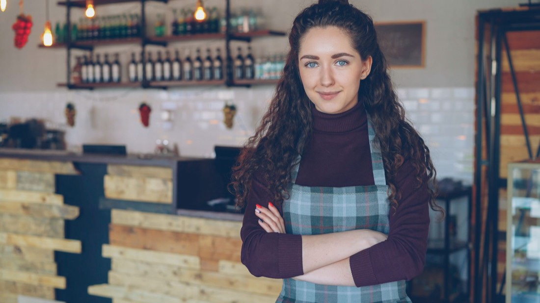 A cafe owner in an apron stands with arms crossed in her coffee shop.