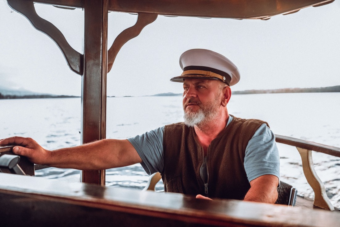 A boat captain in a cap steers from the wheel with open water behind him.