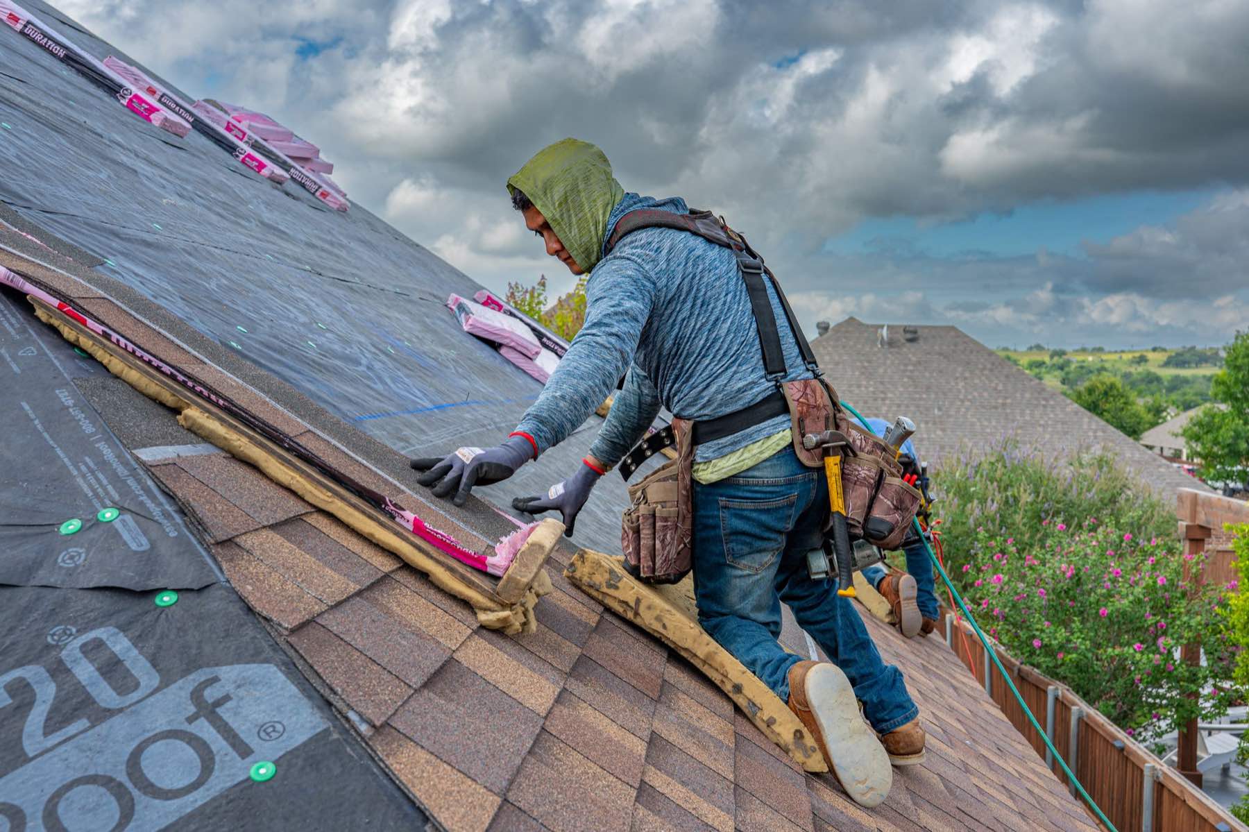 A roofer kneels on a pitched roof, nailing down new shingles under a bright sky.